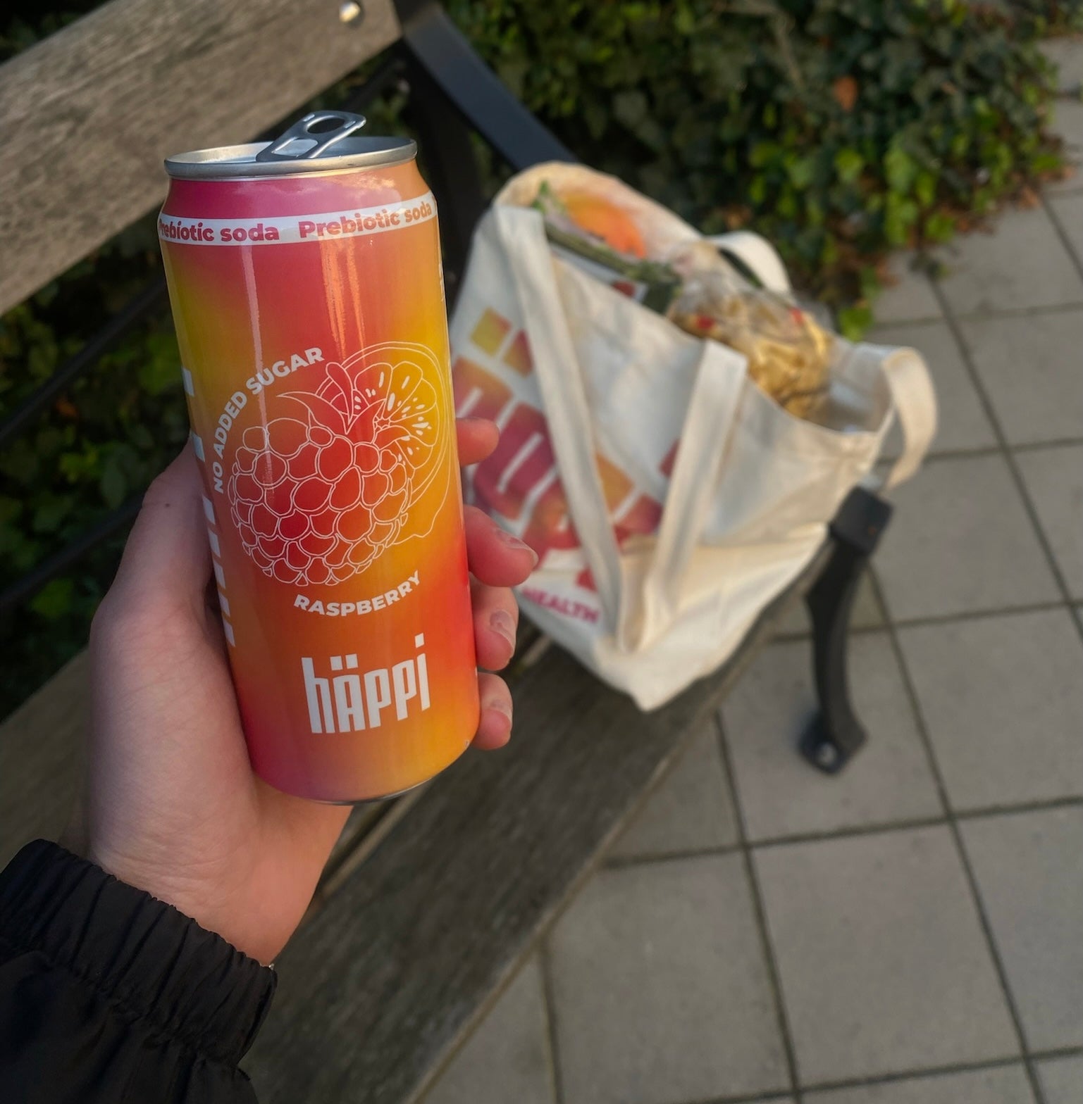 A vibrant can of hÄppi prebiotic soda in raspberry flavor held in hand, showcasing "No Added Sugar" labeling. Positioned outdoors on a park bench, with a reusable tote bag filled with groceries in the background.